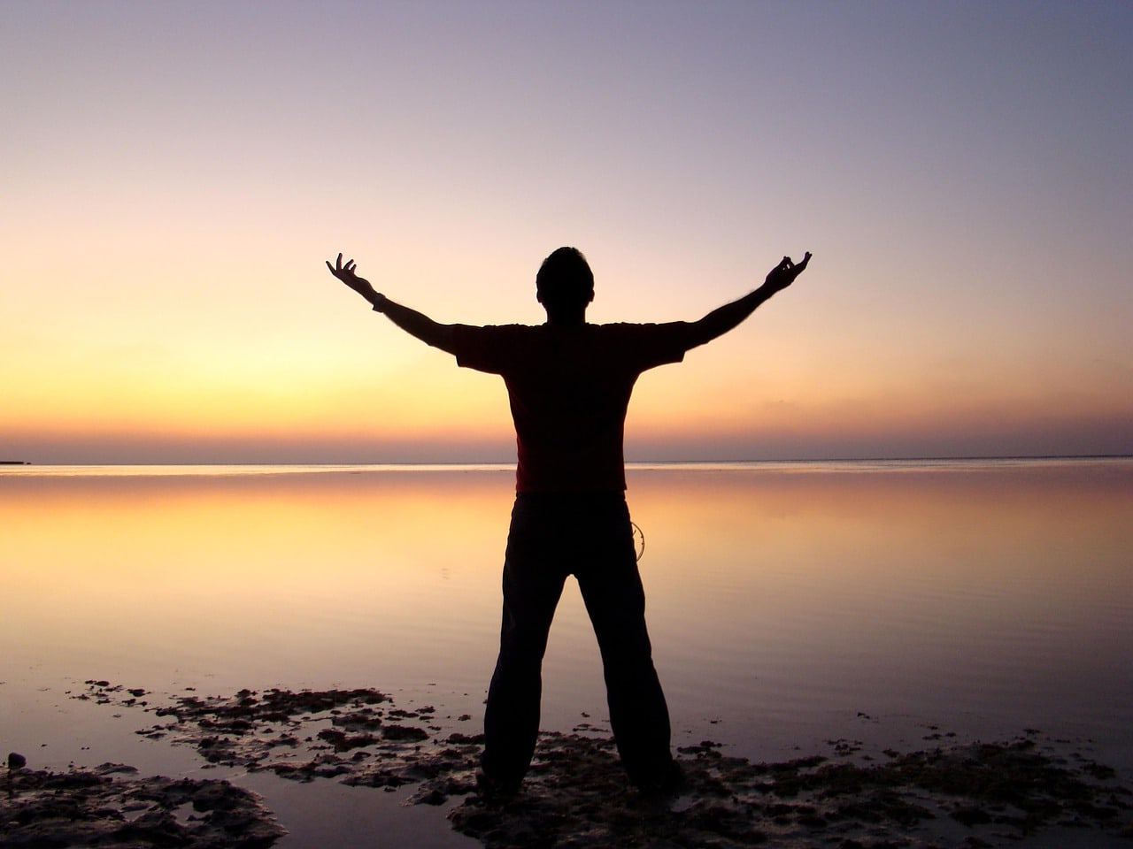A person with outstretched arms silhouetted against a bright sky and calm water